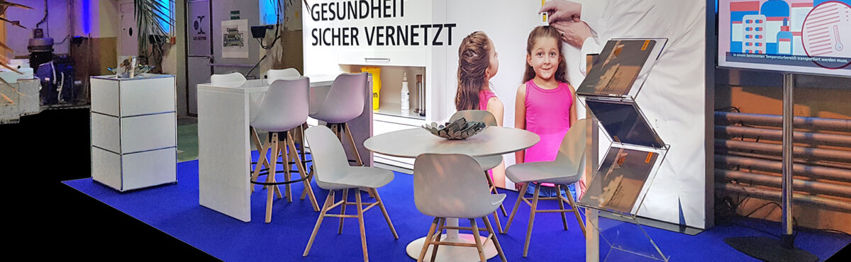 zickzack prospektstaender an einem messestand mit beleuchteter rueckwand sowie tisch und stuehlen